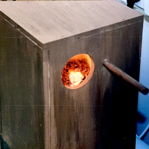 Art work. Wooden bok with peep hole. Through the peephole you see a doll sitting in the corner of the box, with a dribble of blood coming from her mouth.